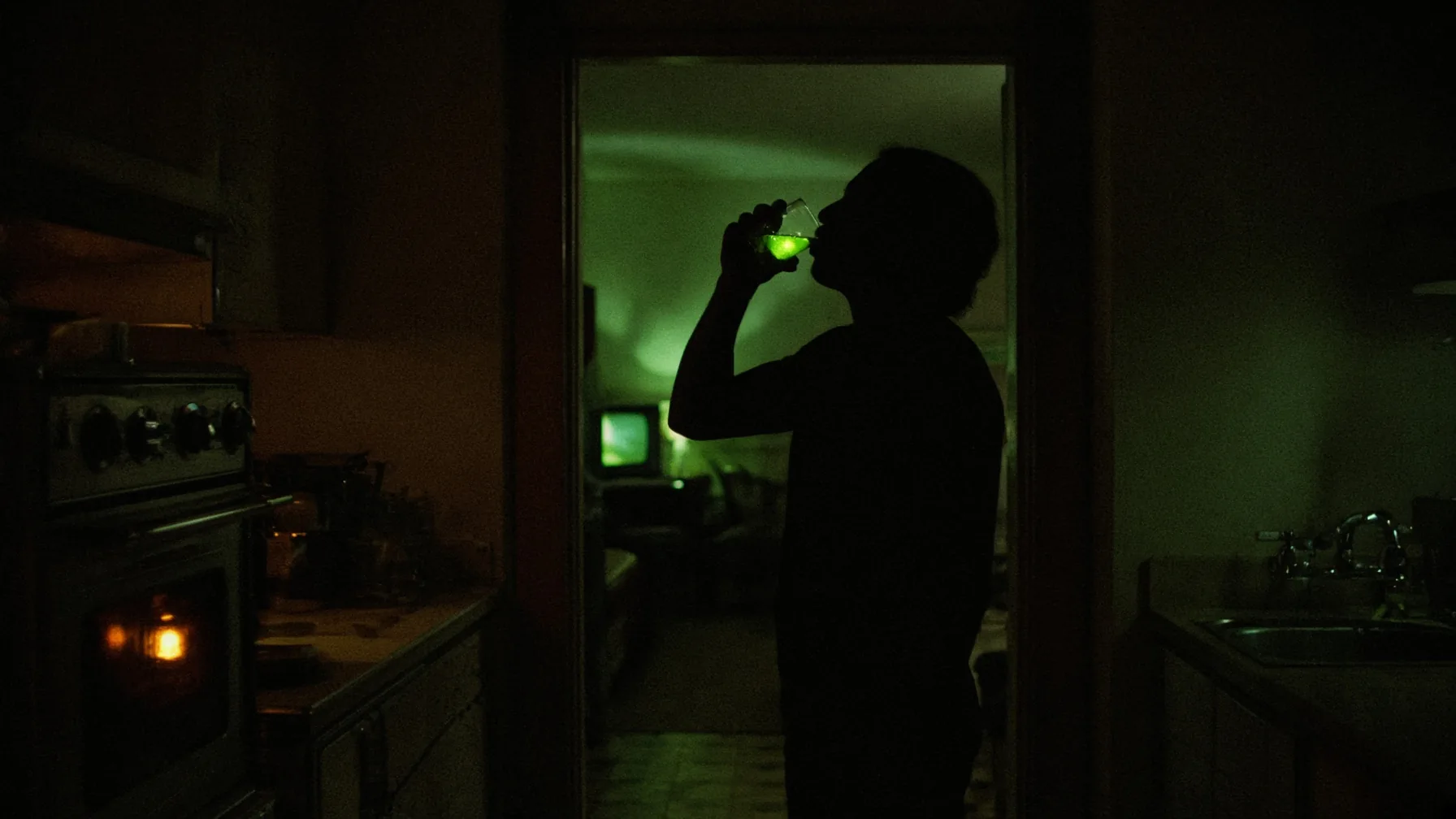A silhouetted person drinks a glass of water that glows green in the light of a television in the next room.